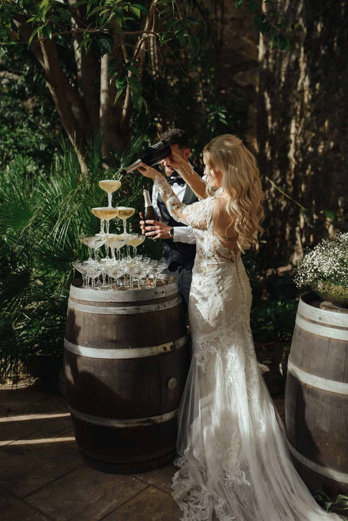 Bride and groom pouring champagne into a coupe glass tower on a wooden barrel in a shaded garden