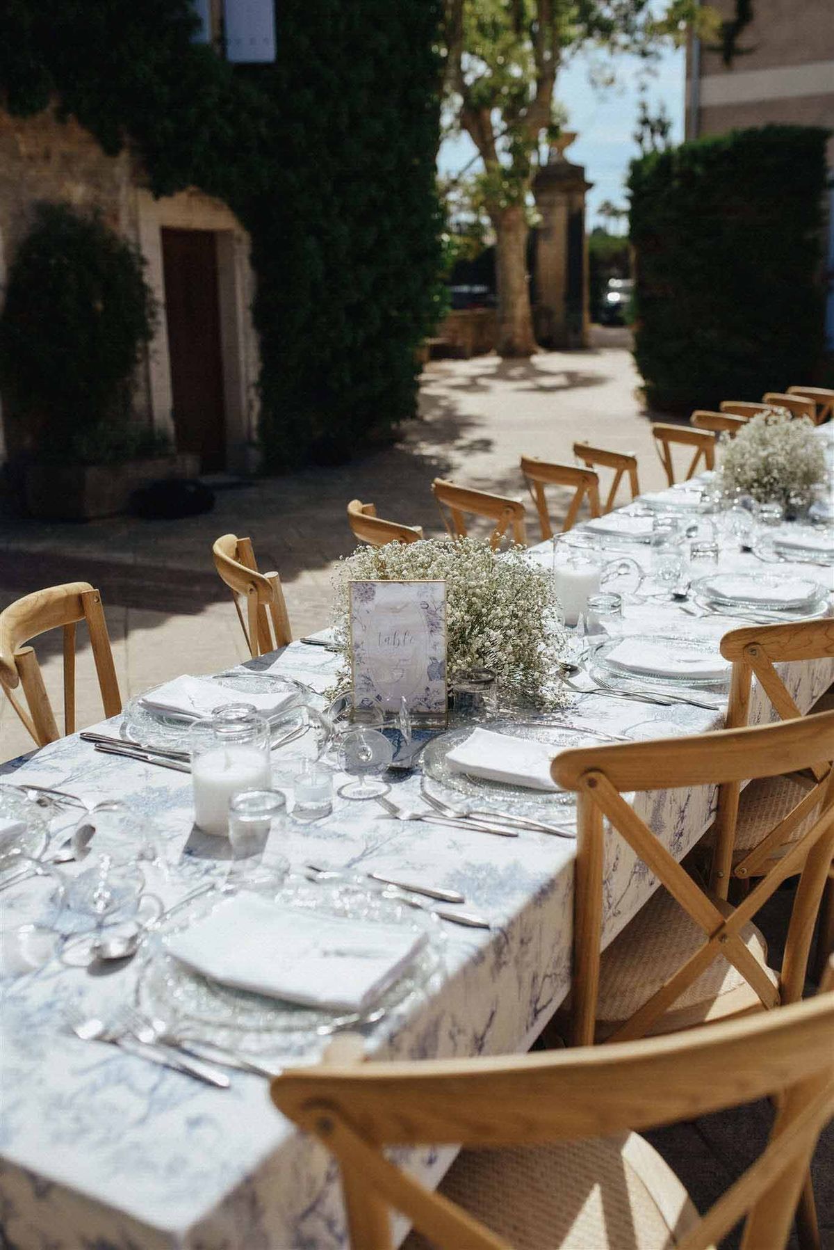 Long banquet table with pale blue linen, white plates, wooden cross-back chairs and baby's breath centerpieces in a stone courtyard.