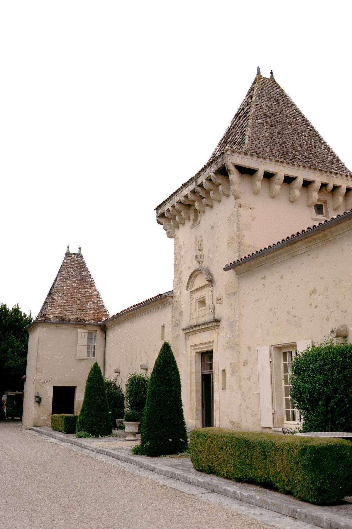 Cream chateau with pointed towers, conical topiary, and box hedging on gravel drive in overcast light