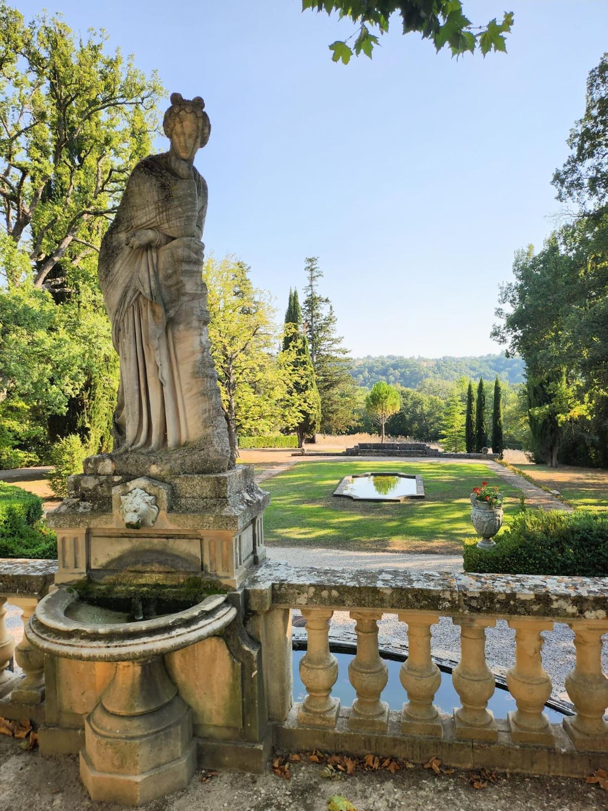 An outdoor shot of a formal French estate garden, captured from an elevated stone terrace. In the foreground, a large classical stone statue of a draped female figure stands atop a pedestal with a lion-head fountain and basin. A stone balustrade runs along the terrace edge, with a long narrow reflecting pool below. The garden extends into a manicured lawn with a small octagonal mirror pool at its center, flanked by a stone urn planted with red geraniums. Tall Italian cypress trees frame a central axis leading to a wooded hillside in the distance. No people are present in the image. Wide shot, no wedding-specific decor visible. Potential venue feature image.