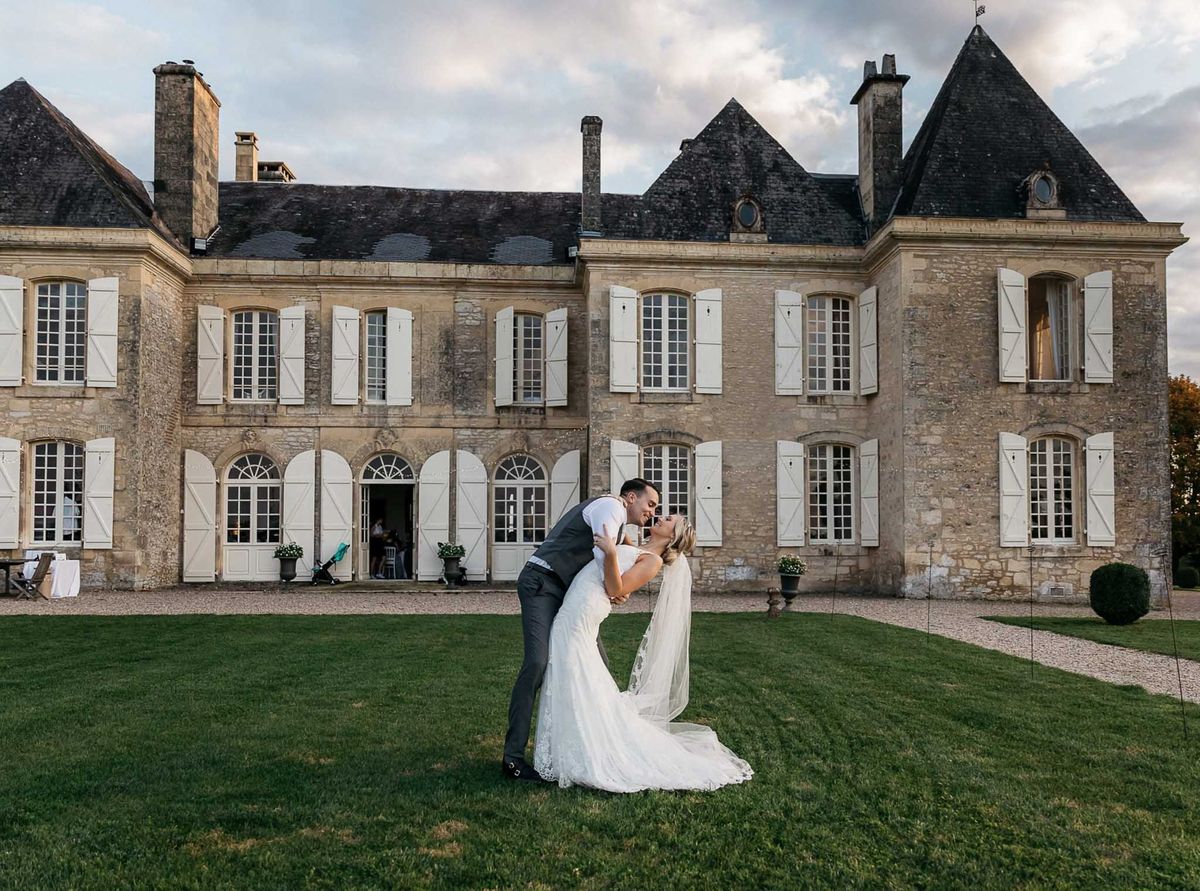 Groom dipping bride in ivory lace gown with cathedral veil before limestone chateau at golden hour