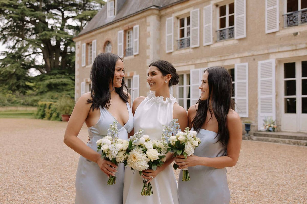 Bride in white gown and two bridesmaids in pale blue sleeveless gowns holding bouquets in front of a French château facade.