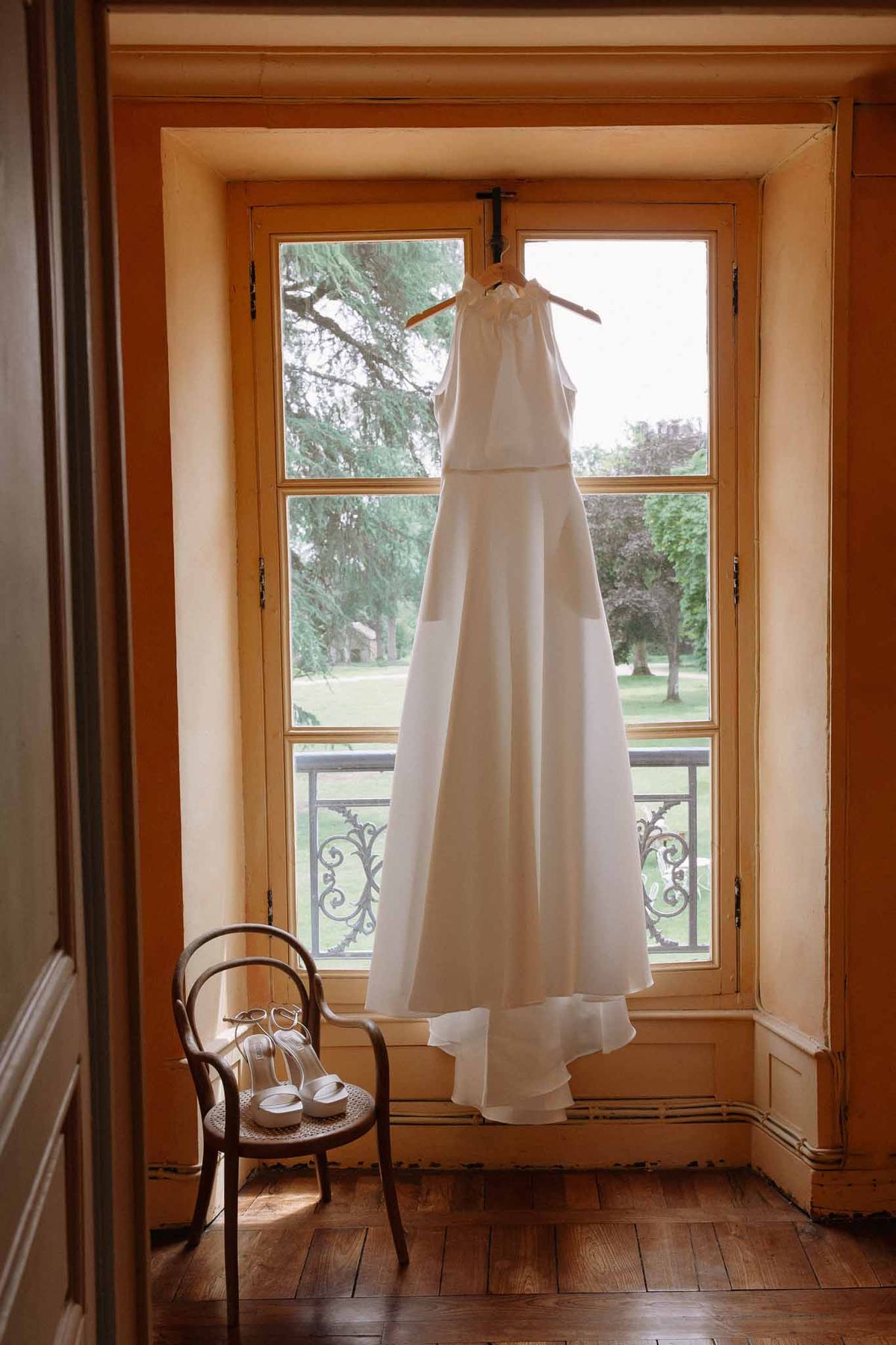 Ivory halter wedding dress hanging from wooden hanger in front of tall French doors in a paneled chateau room