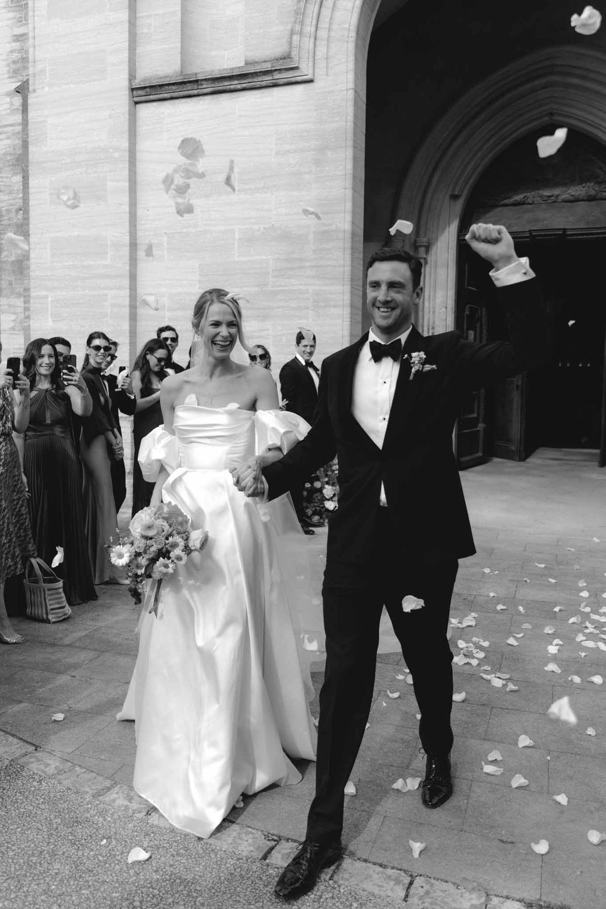 Black-and-white photo of couple exiting stone church with petal toss and guests lining the arched doorway