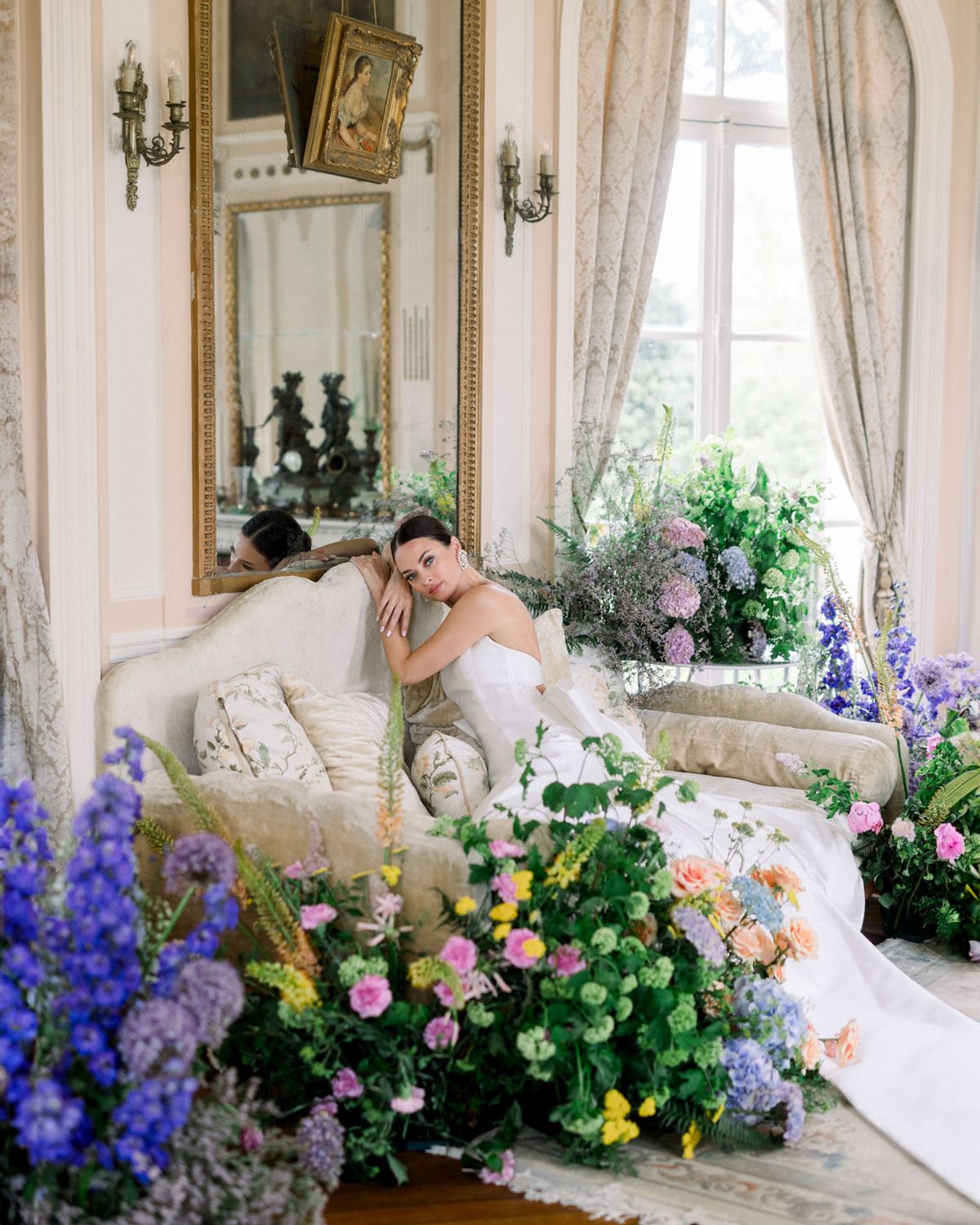 Bride on ivory velvet sofa surrounded by maximalist blue pink and peach floral installations in salon