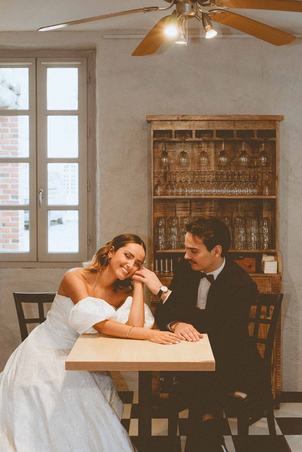 Bride and groom seated at bistro table smiling at each other with rustic wine bar cabinet behind them