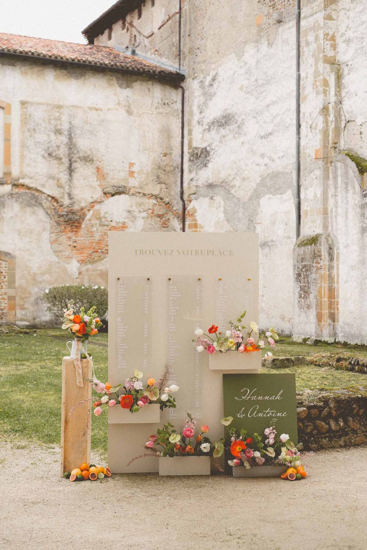 A wedding seating chart display set up outdoors in front of a historic stone building with exposed brick and weathered plaster walls. The display consists of a large cream/beige rectangular board headed with the text 'TROUVEZ VOTRE PLACE' in olive green lettering, with translucent acrylic panels hanging from gold pins listing guest names in white calligraphy. A dark olive green sign reading 'Hannah & Antoine' in white script is positioned at the lower right. The display is decorated with multiple small cream planter boxes holding an arrangement of colorful blooms including orange and red poppies, pink and white ranunculus, orange tulips, pink stock flowers, and anthurium leaves. Sliced citrus fruits — blood oranges and limes — are scattered at the base of the display alongside a wooden pedestal holding a matching floral arrangement. The overall color palette combines cream, olive green, and vivid warm-toned florals in orange, red, coral, and pink. Wide shot, slightly angled, capturing the full display in context with the architectural backdrop.