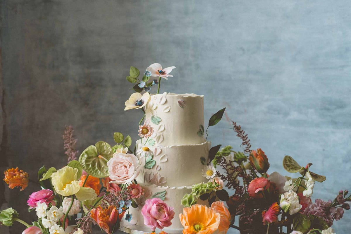 A close-up detail shot of a three-tier wedding cake with an ivory textured buttercream finish, decorated with handcrafted sugar flowers including white anemones, blush daisy-like blooms, pale yellow flowers, and green sugar leaves cascading across the tiers. The cake is surrounded at its base by a loose, abundant arrangement of fresh flowers in a vibrant mixed palette — orange poppies, hot pink ranunculus, blush garden roses, yellow parrot tulips, orange tulips, white narcissus, blue forget-me-nots, green anthurium leaves, and burgundy astilbe. The background is a muted blue-grey painted or textured wall, giving the composition a studio or styled editorial feel. The overall floral styling is bold and colorful with a garden-gathered aesthetic, contrasting with the pale, delicate cake decoration.