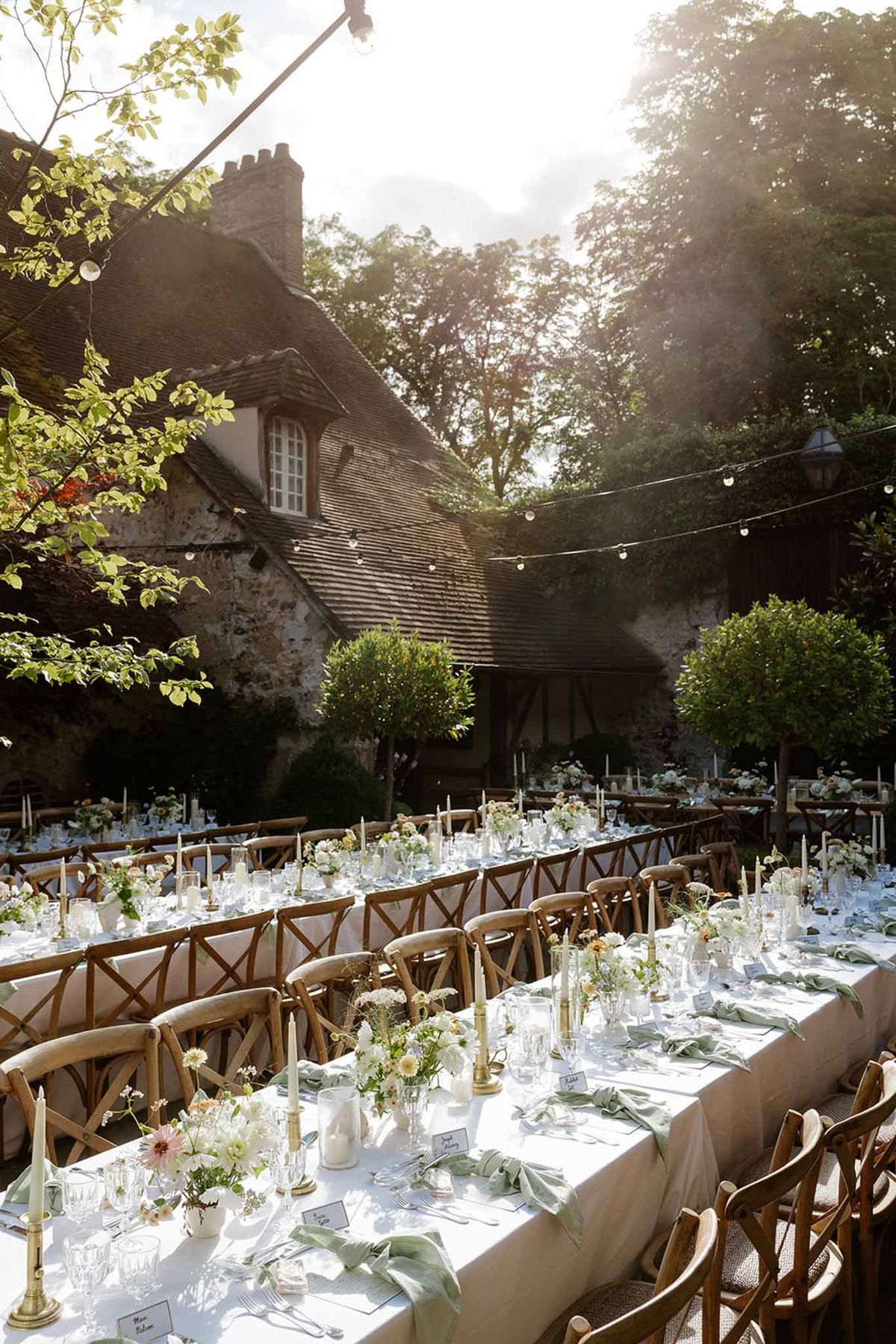 Outdoor courtyard reception with long tables, brass candlesticks, and string lights by stone manor