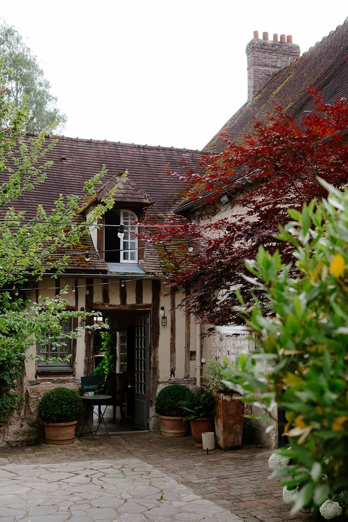 Timber-framed cottage with clay tile roof and brick chimneys in a courtyard with Japanese maple and ivy