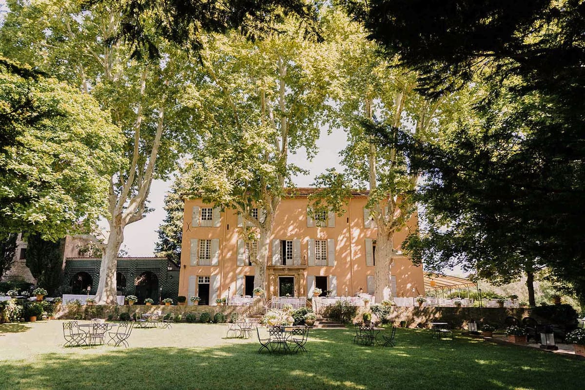 Ochre Provencal manor with grey-green shutters, white reception tables on terrace, and bistro chairs on lawn