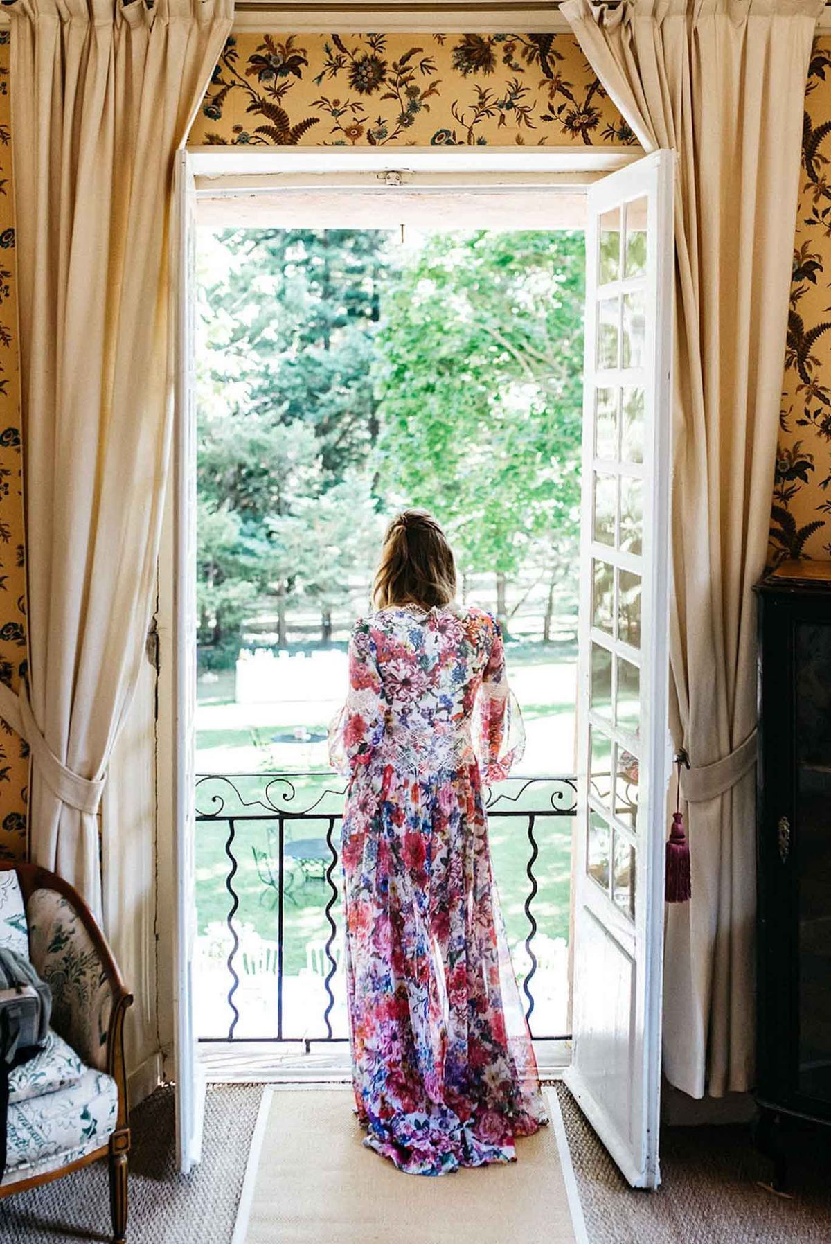 Bride in floral printed robe standing in open French doors of a chateau room with ornate gold wallpaper