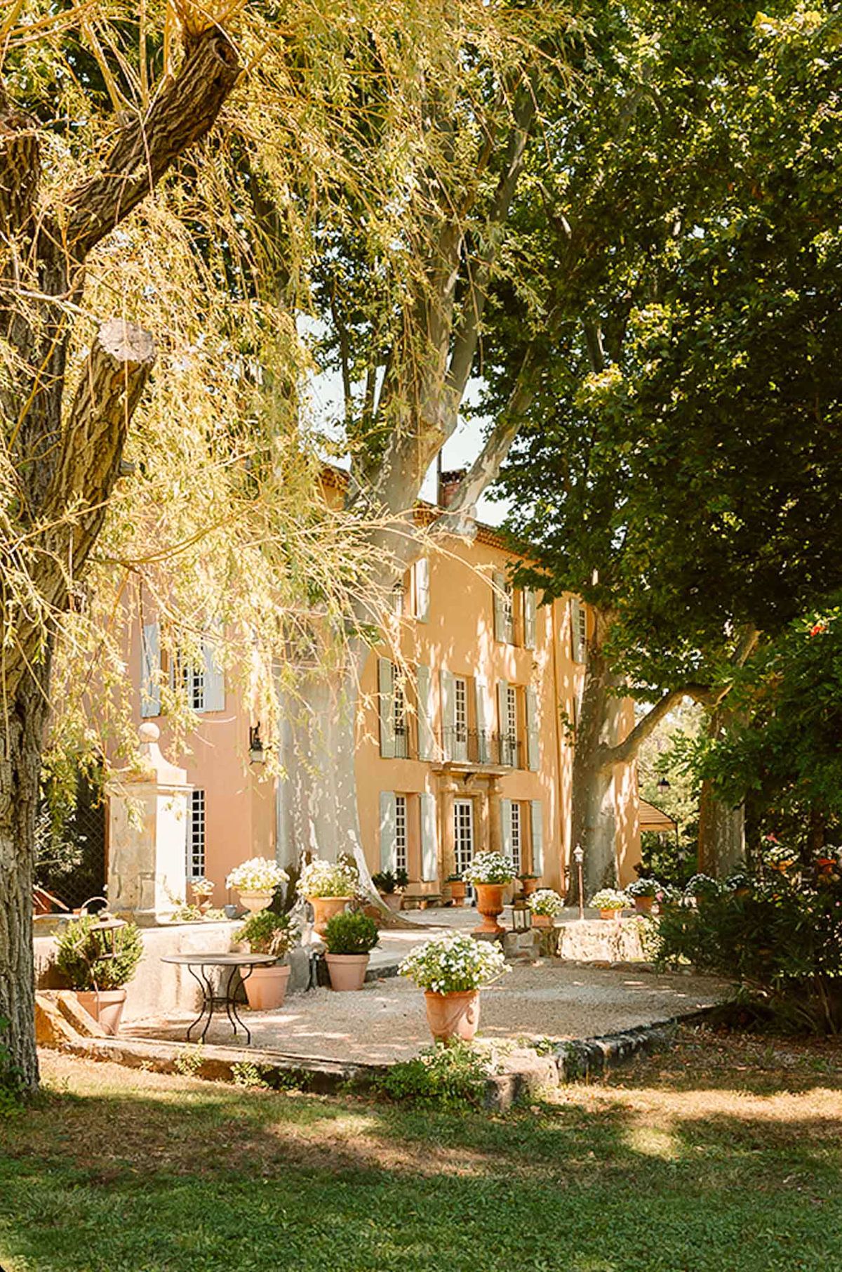 An outdoor wide shot of a two-storey Provençal mas or bastide-style manor house with warm terracotta-peach rendered walls and pale sage-green shutters. The entrance forecourt features a gravel courtyard lined with large terracotta urns planted with white flowering arrangements, clipped boxwood spheres, and additional potted greenery leading up to the front steps and entrance door. A wrought-iron bistro table and chairs are visible to the left near a stone fountain or well feature. No people are present in the image. The styling is classic French Provençal with a warm, sun-lit palette of terracotta, sage, and white. Potential venue feature image.