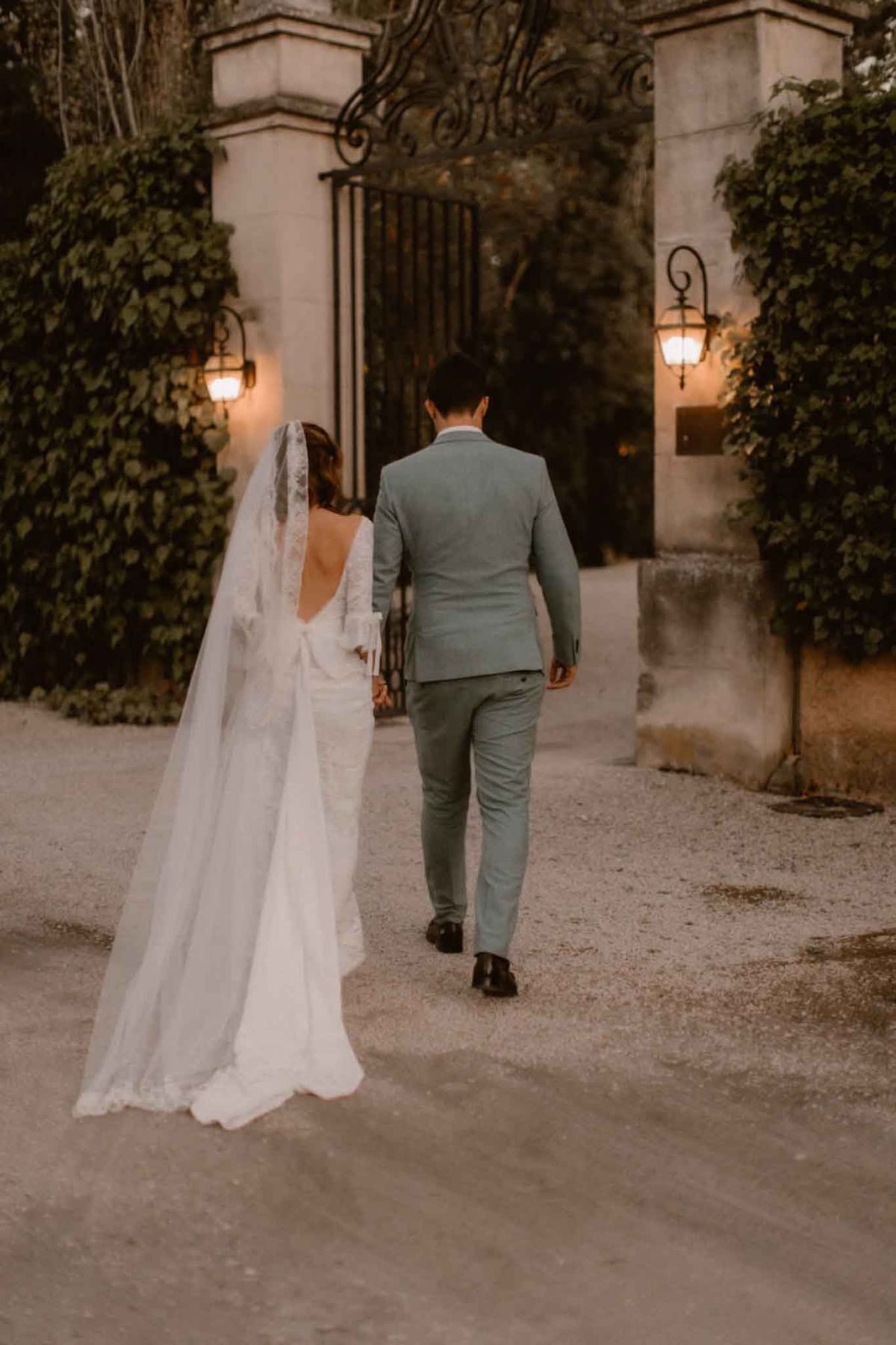 Couple walking from behind toward wrought iron gate at dusk bride in lace gown groom in sage green suit