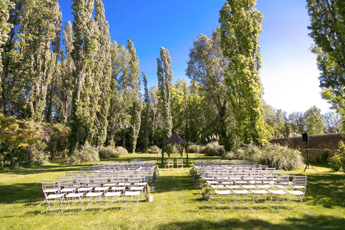 An outdoor ceremony setup photographed in a wide shot on a large lawn enclosed by a stone wall, with tall poplar trees forming a natural backdrop. Rows of white folding chairs are arranged in two sections divided by a central aisle, accommodating approximately 80–100 guests. At the far end of the aisle, a small dark brown canopy or pergola structure serves as the ceremony focal point, flanked by two yellow chairs, likely for the couple. Small potted plants in white pots mark the ends of the aisle rows, and a PA speaker on a stand is visible to the right. The overall decor style is simple and understated, with a neutral white chair palette and minimal floral decoration. Potential venue feature image.