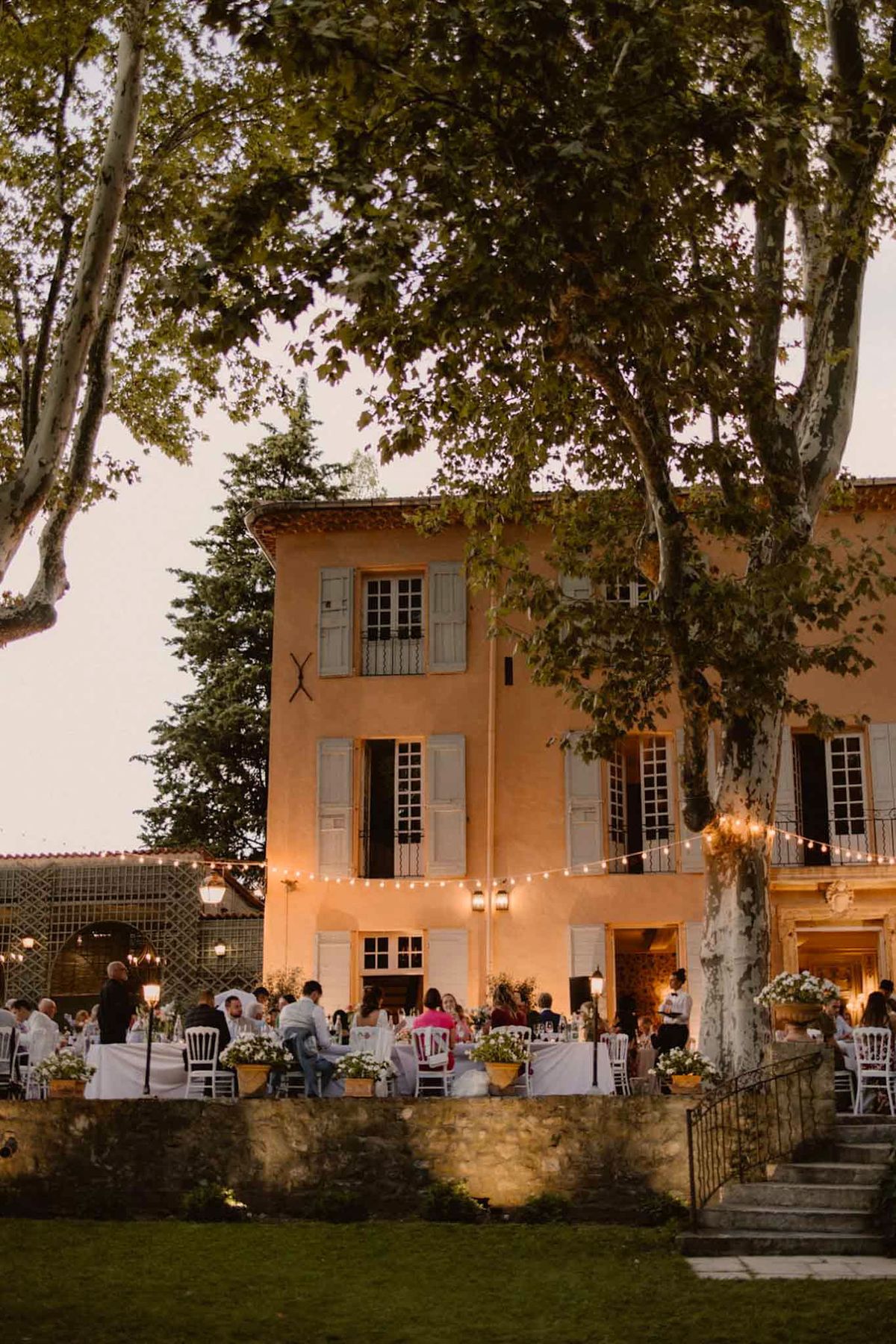 Outdoor wedding reception on Provencal bastide terrace with round tables and festoon lights at dusk