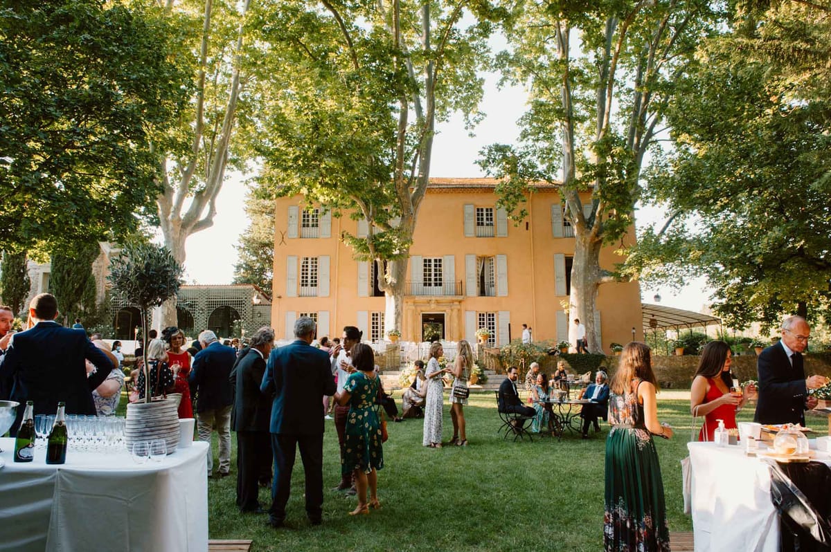 Guests mingling on lawn during cocktail hour at ochre Provencal bastide with champagne bar tables at golden hour