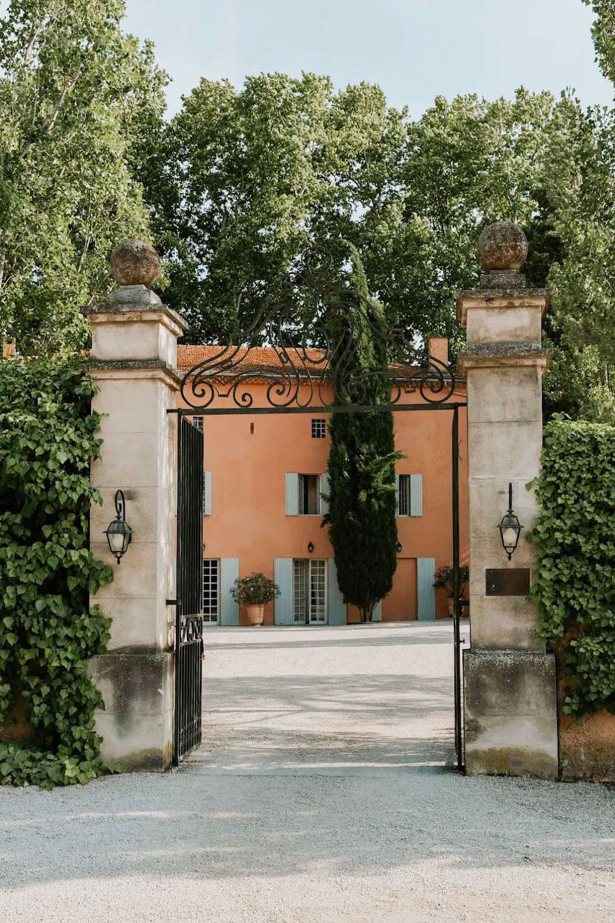 A wide shot of a French country estate viewed through an open ornamental wrought-iron gate flanked by two stone pillars topped with carved stone ball finials, each fitted with a black lantern-style wall light. The gate opens onto a gravel courtyard leading to a two-story mas or bastide-style manor house with terracotta-orange rendered walls, a red-tiled roof, and pale blue-grey shutters on all windows and doors. A tall cypress tree stands in front of the building, and terracotta pots with flowering plants flank the entrance. No people are visible in the image. The composition is a straight-on wide shot framed symmetrically by the gateposts. Potential venue feature image.