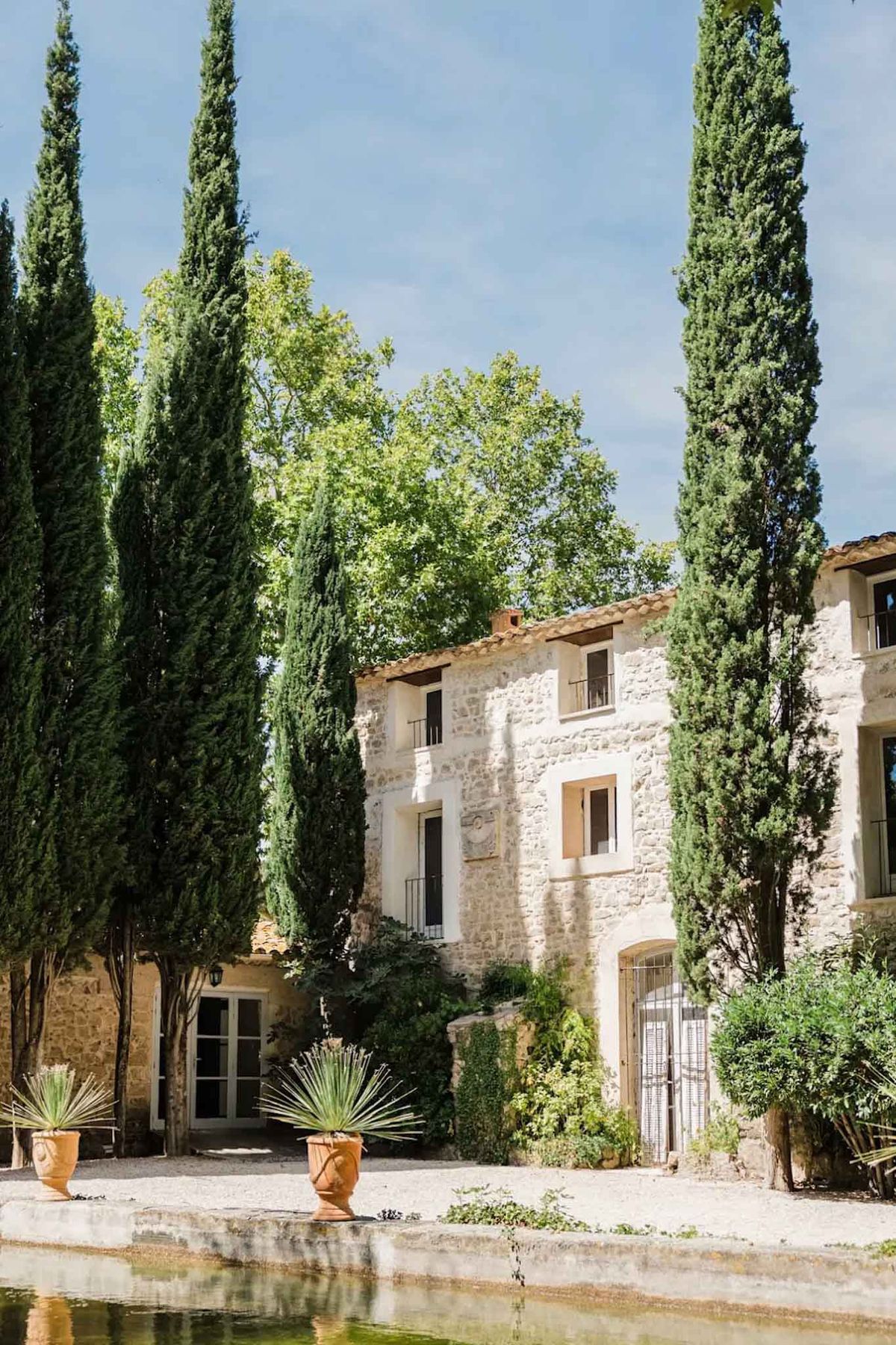 An outdoor wide shot of a traditional Provençal mas-style property with exposed limestone block façades, terracotta roof tiles, and wrought iron balconies and gate details. The two-storey main building is flanked by tall Italian cypress trees and features French doors on the ground level and shuttered windows with warm ochre surrounds. Two terracotta urn planters holding spiky yucca-style plants are positioned on the gravel courtyard in the foreground, with a long rectangular ornamental stone basin reflecting the scene in the immediate foreground. The grounds include dense established greenery and shrubs around the building's base. Potential venue feature image.