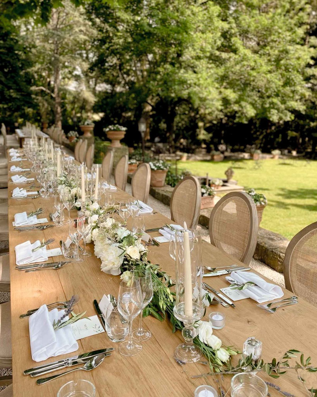 Long farm table set for outdoor reception with white peony runner, taper candles, and rattan chairs in Provence garden