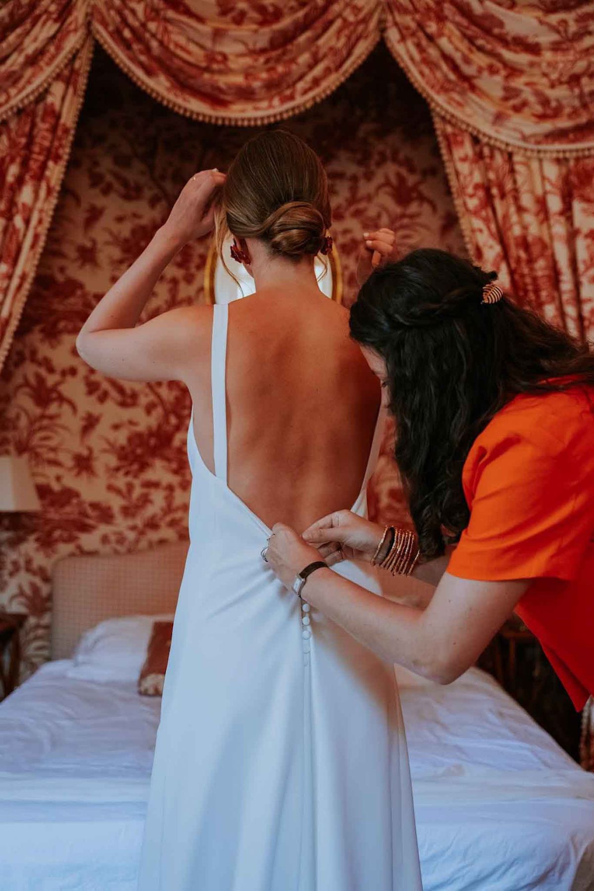 A getting-ready scene photographed indoors in a bedroom, showing the bride from behind as a woman in an orange top fastens the row of small covered buttons running down the back of her white wedding dress. The bride's dress features a deep open back with thin cross-back straps and a minimalist, sleek silhouette. Her hair is styled in a low chignon, and she wears deep red drop earrings. The helper, who has dark curly hair and is wearing multiple gold and red bangles, leans in to work the buttons. The room is decorated in classic toile de Jouy fabric in red and cream, visible on the canopy and draping of a four-poster bed, giving the setting a traditional French interior style. A white duvet is visible on the bed in the foreground. The shot is a medium close-up portrait taken from behind.