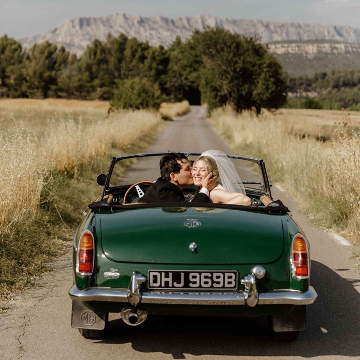 Couple kissing in vintage green MG convertible on rural Provence road with limestone massif in background