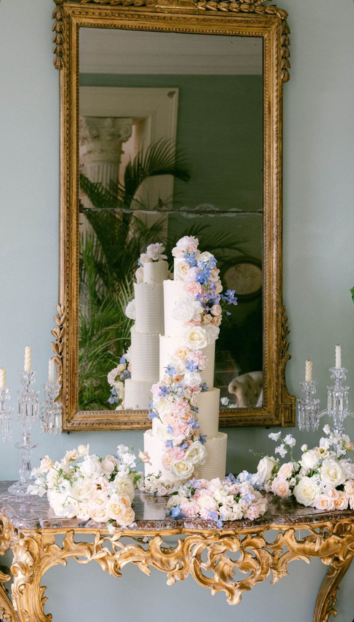Five-tier ribbed buttercream wedding cake with cascading pink roses and blue delphinium on a gilt console table