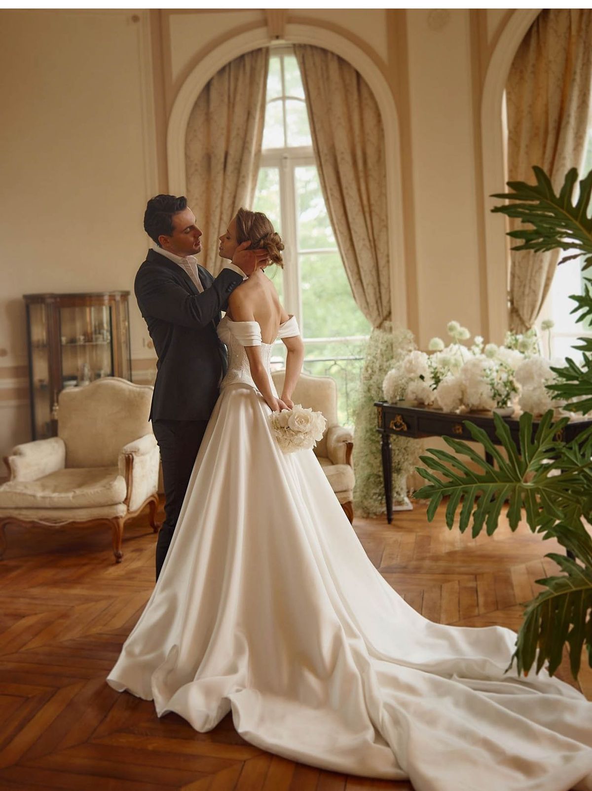 A couple portrait taken indoors in what appears to be a formal salon or reception room of a French château, featuring herringbone parquet flooring, warm cream-painted walls, and tall arched windows dressed with gold-toned damask curtains. The groom, wearing a dark navy suit with a white dress shirt, gently cups the bride's face as they share an intimate moment. The bride wears an off-the-shoulder ivory satin ball gown with a structured bodice and an extended cathedral-length train pooling across the floor, and she holds a compact bouquet of cream roses; her hair is styled in a low chignon. In the background, a black antique console table is heavily decorated with large arrangements of white peonies, hydrangeas, and roses in a classic, all-white floral palette, accompanied by large tropical foliage plants on either side. The overall styling is classic and formal, with the room's Louis XV-style armchairs and glass display cabinet reinforcing the period interior setting. The composition is a full-length portrait shot from slightly behind and to the side of the couple.