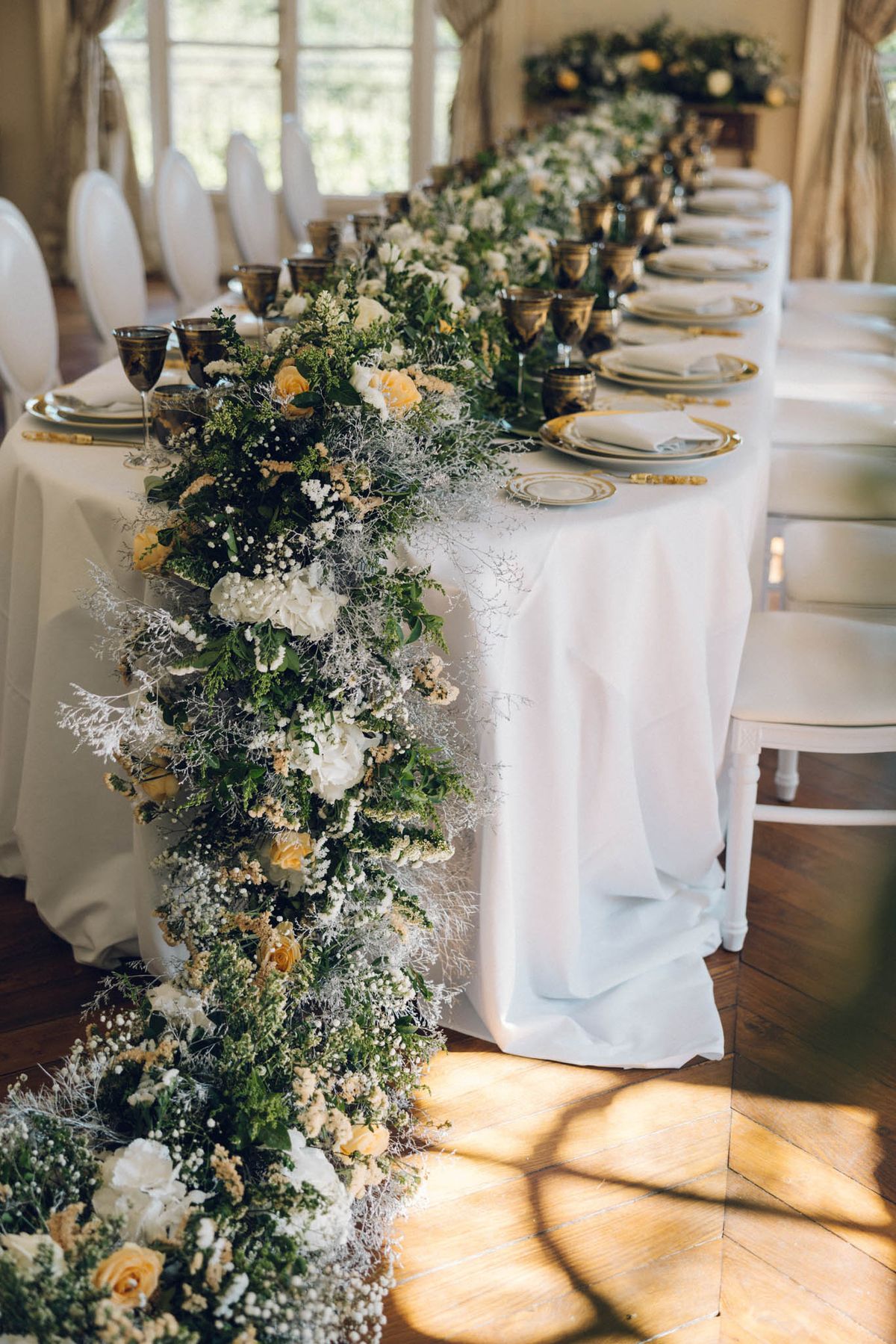 Detail shot of a long rectangular reception table set inside a bright, windowed room with parquet flooring and draped curtains. The table is covered in a white linen cloth and features an abundant floral garland runner that cascades off the end, composed of lush greenery, white hydrangeas, ivory baby's breath, soft yellow roses, and wispy silver-grey dried filler stems. Place settings include gold-rimmed charger plates with white dinner plates, folded white napkins, gold flatware, and dark amber-toned metallic goblets. White banquet chairs with curved backs line both sides of the table, and a floral arrangement is visible on a sideboard in the background. The overall decor palette is white, gold, soft yellow, and green with a classic, garden-inspired style.