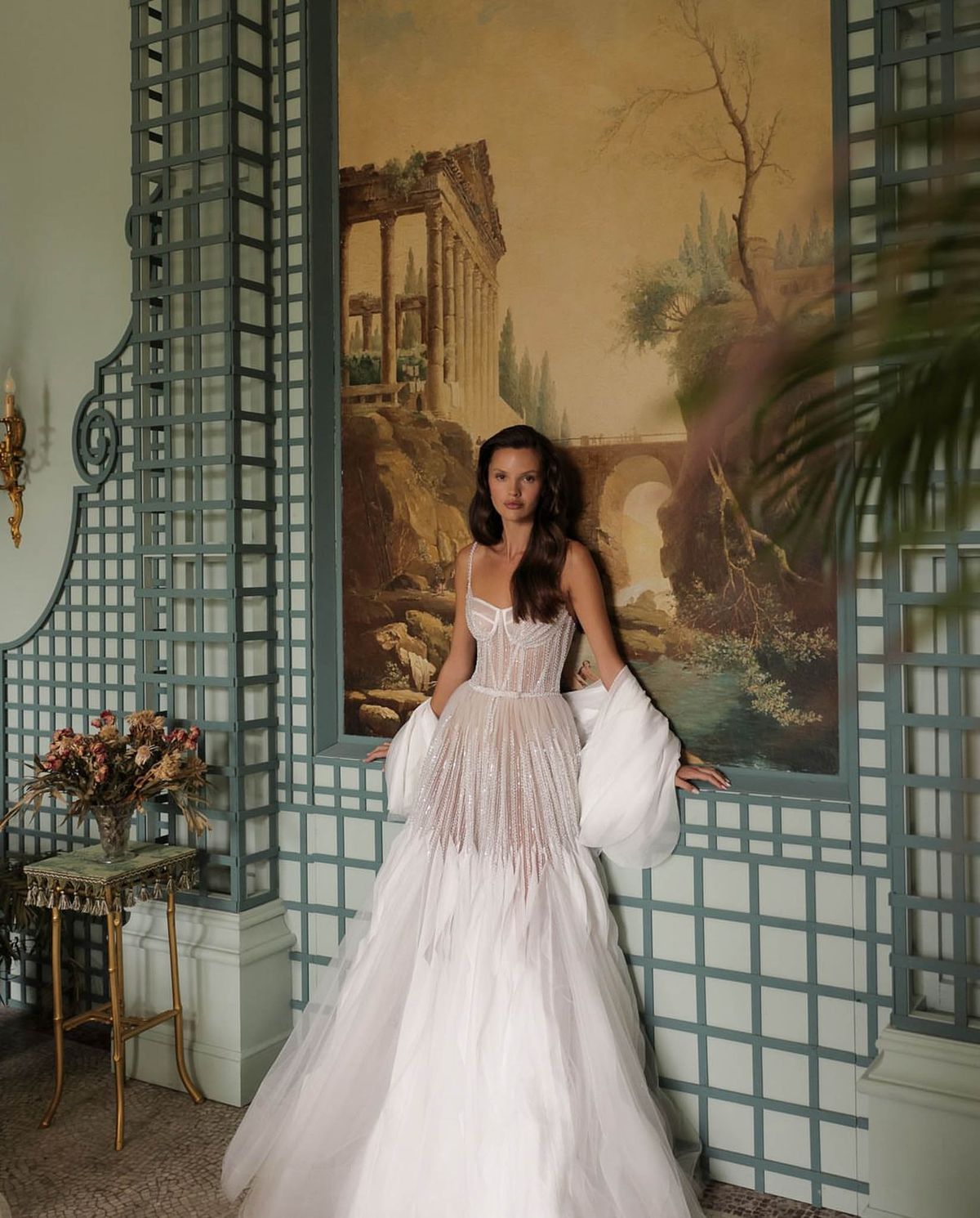 A bridal portrait shot indoors featuring one woman in a white ball gown with a structured corset bodice heavily embellished with crystal or beaded detailing in a radiating pattern, transitioning into a full tulle skirt. She wears a voluminous white wrap or stole draped over her arms and has long, loosely waved dark hair. The setting is a formal interior room featuring blue-grey trellis-style wall paneling, a large classical landscape mural depicting ruins and a bridge, a gold ornate side table holding a dried floral arrangement in muted terracotta and blush tones, and a wall-mounted candle sconce to the left. The composition is a medium full-length portrait with the subject leaning slightly against the trellis partition. The overall styling aesthetic is classic and formal with ornate French interior details.