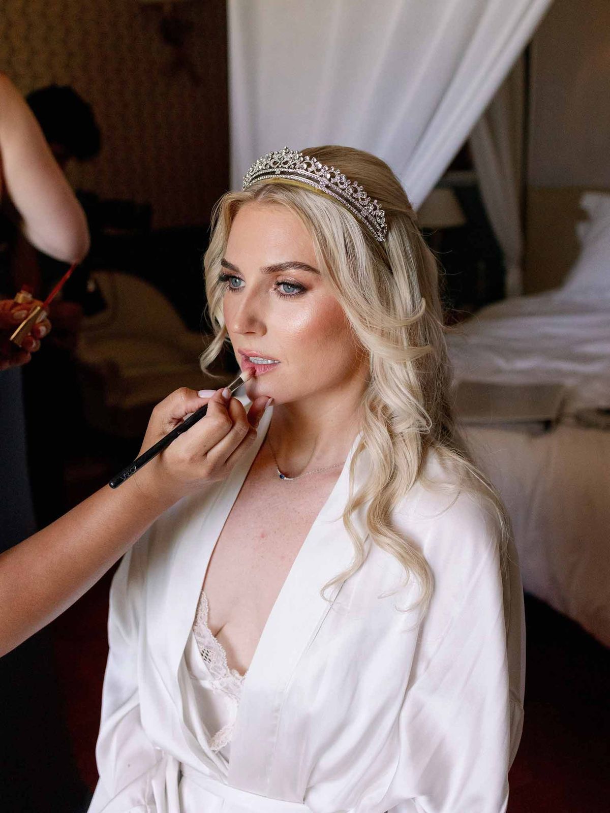Bride in white silk robe and crystal tiara having lipstick applied by makeup artist during getting ready