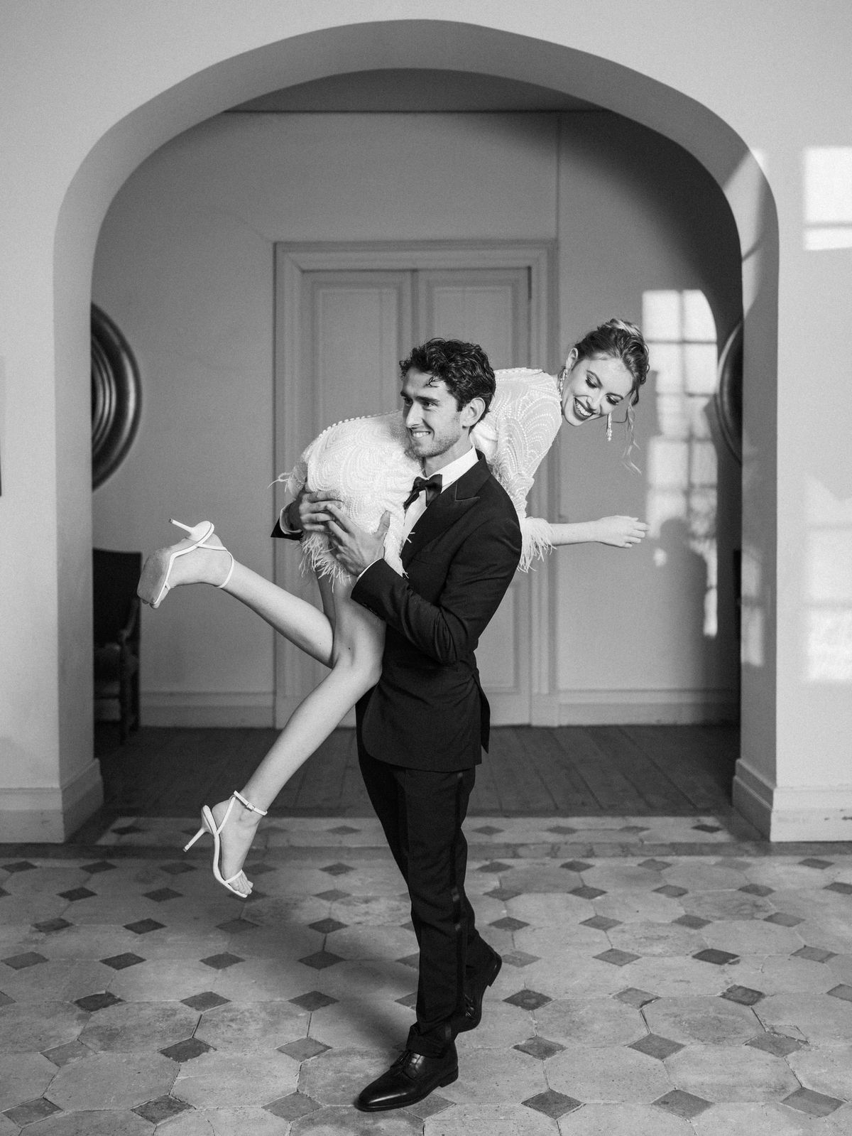 Bride and groom in black and white, groom lifting bride in a classical hallway with diamond-patterned floor