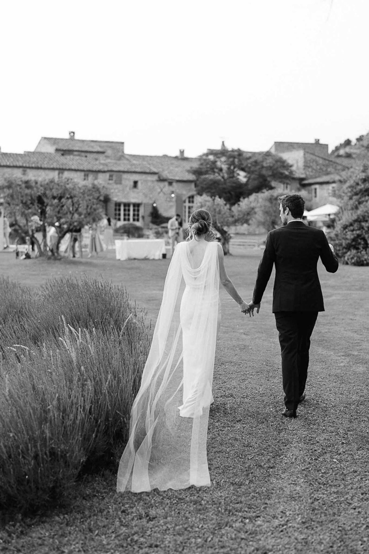 Black and white couple walking hand-in-hand on gravel path of Tuscan estate with olive trees and lavender borders