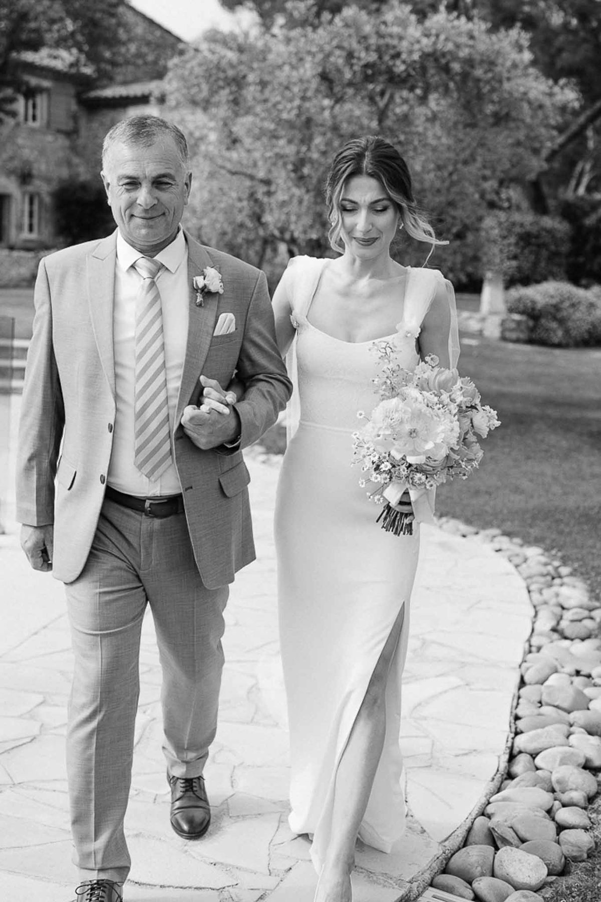 Bride walking arm-in-arm with her father along a garden pathway toward a stone building in a classic outdoor ceremony setting.