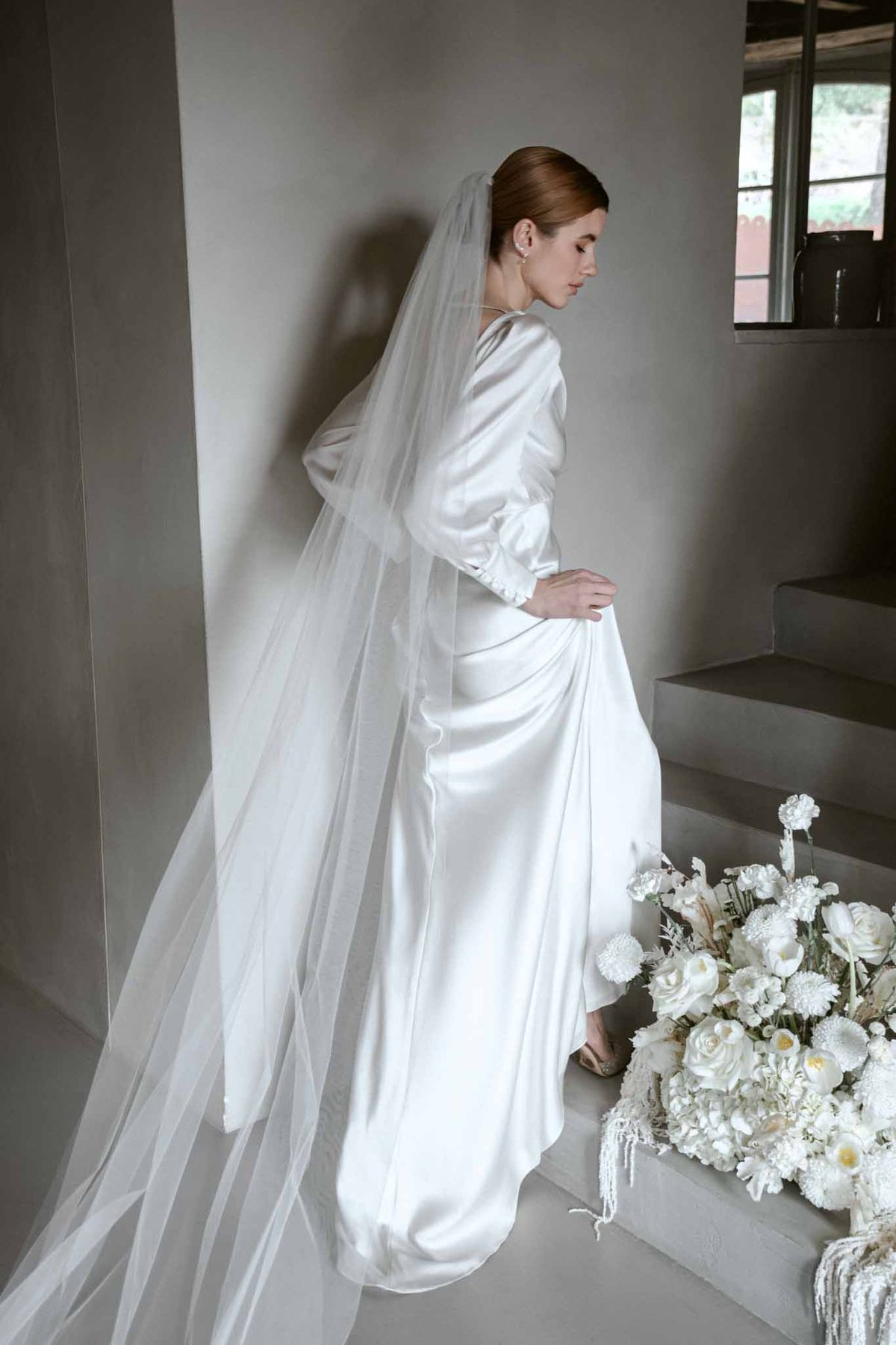 Bride in profile wearing ivory satin gown with long tulle veil and white rose bouquet