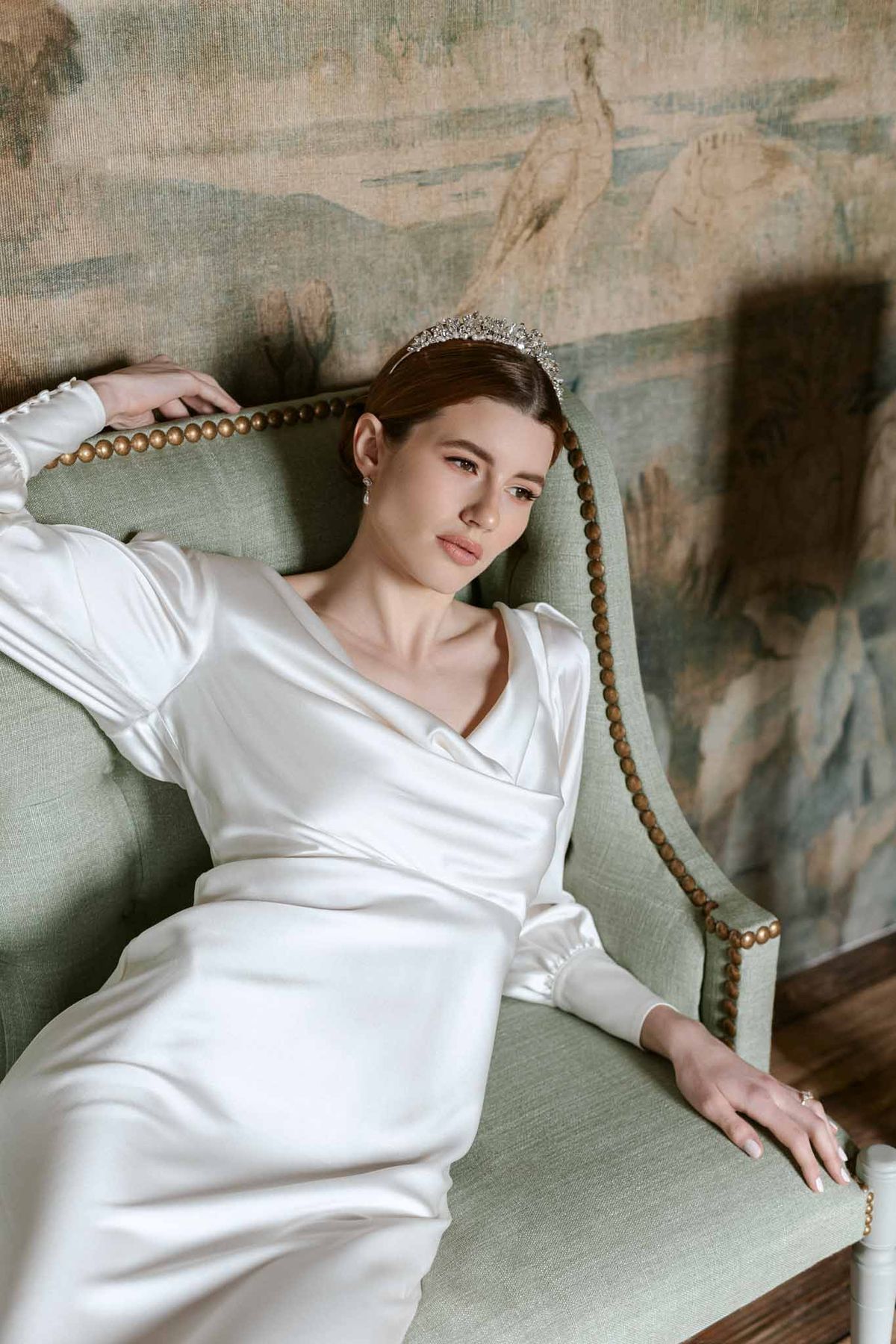 Bride in ivory satin dress with crystal tiara seated on sage green chair by classical mural