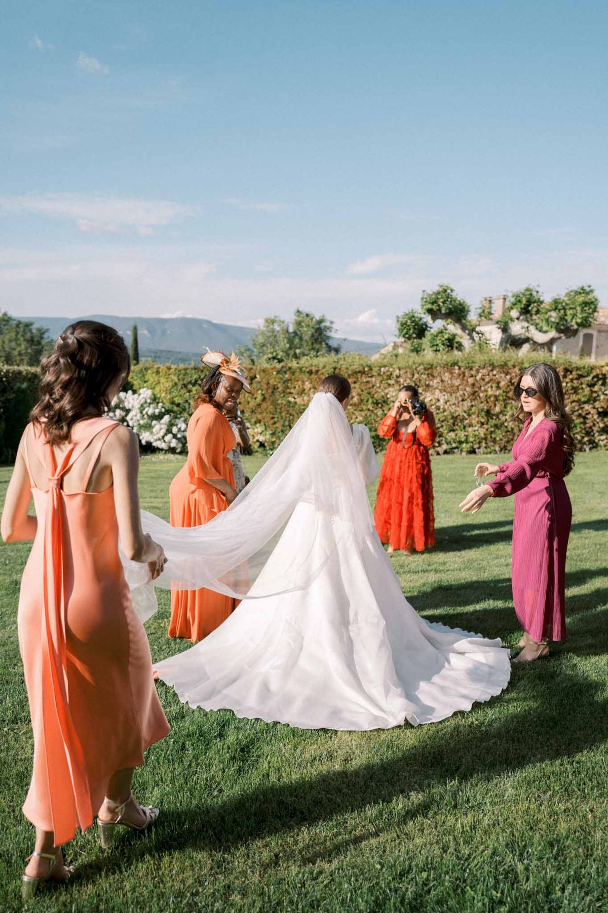 Four women in jewel-toned dresses arrange bride's cathedral veil and train on garden lawn before ceremony