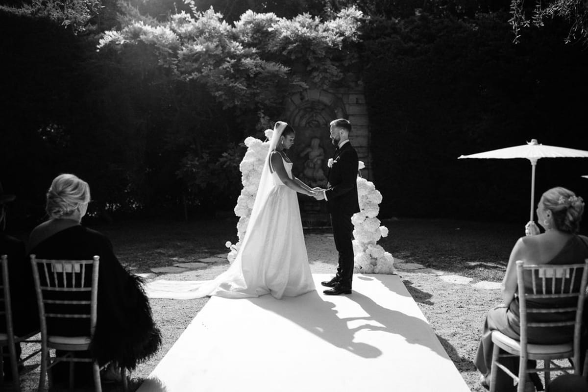 Bride and groom exchanging vows under floral arch at outdoor garden wedding ceremony with guests seated on both sides