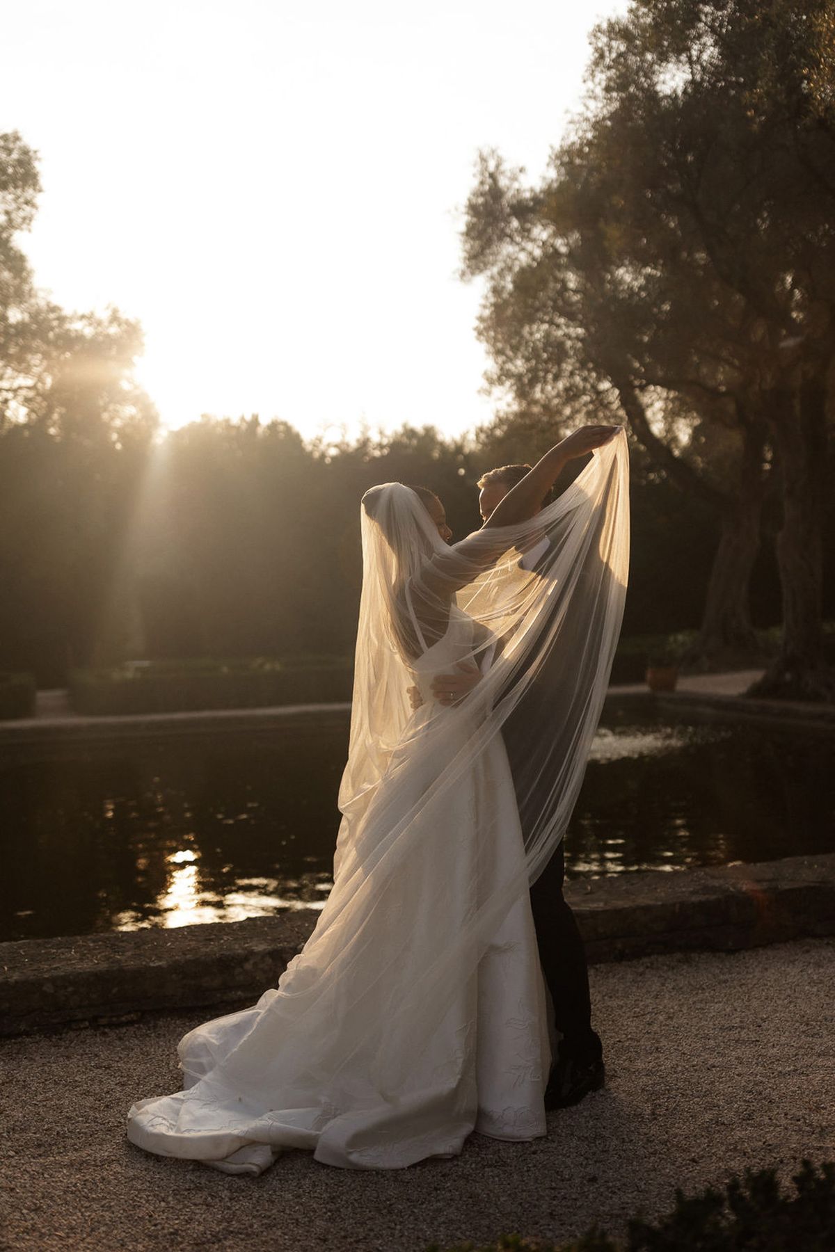 Bride and groom portrait by lake during golden hour with flowing veil in outdoor garden setting