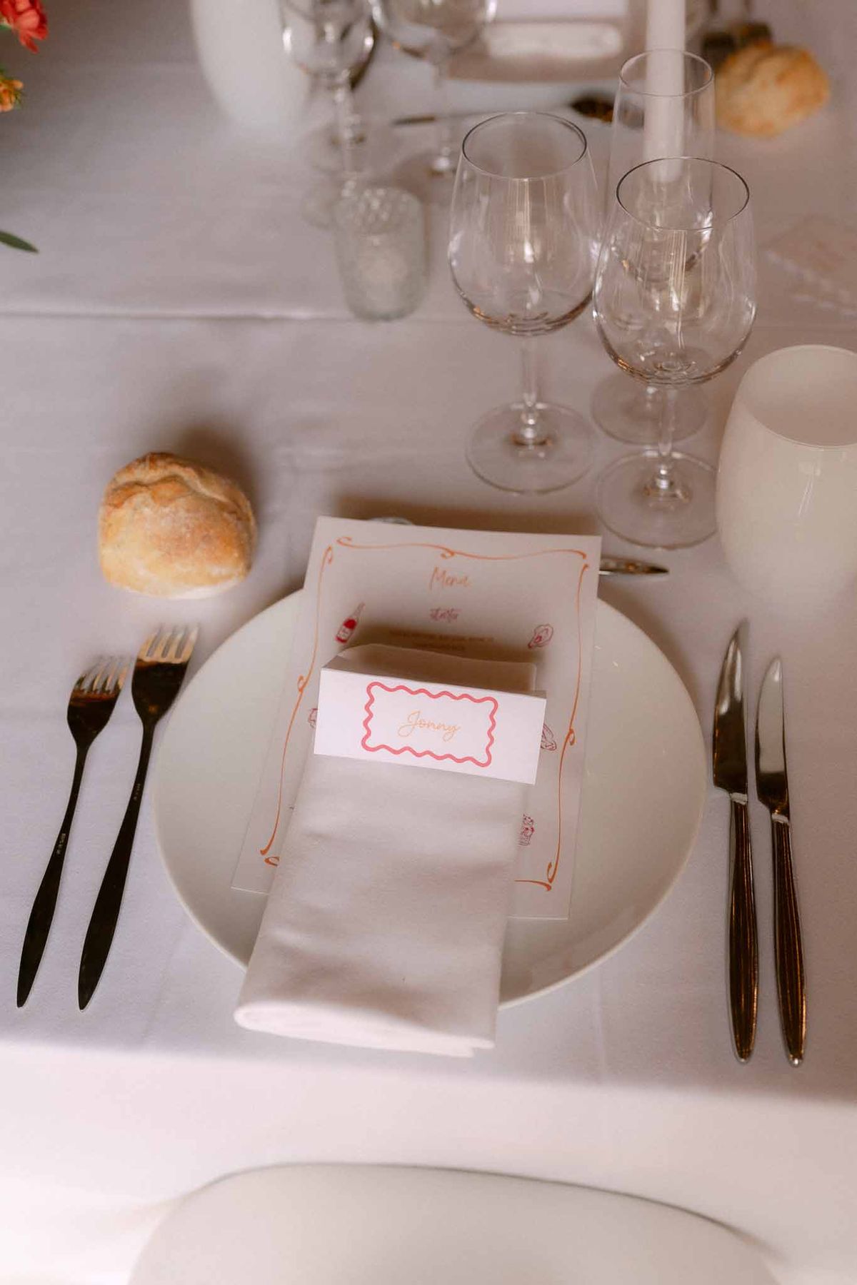 Wedding place setting with white plate, printed menu card with orange border, place card, black matte cutlery, and crystal...