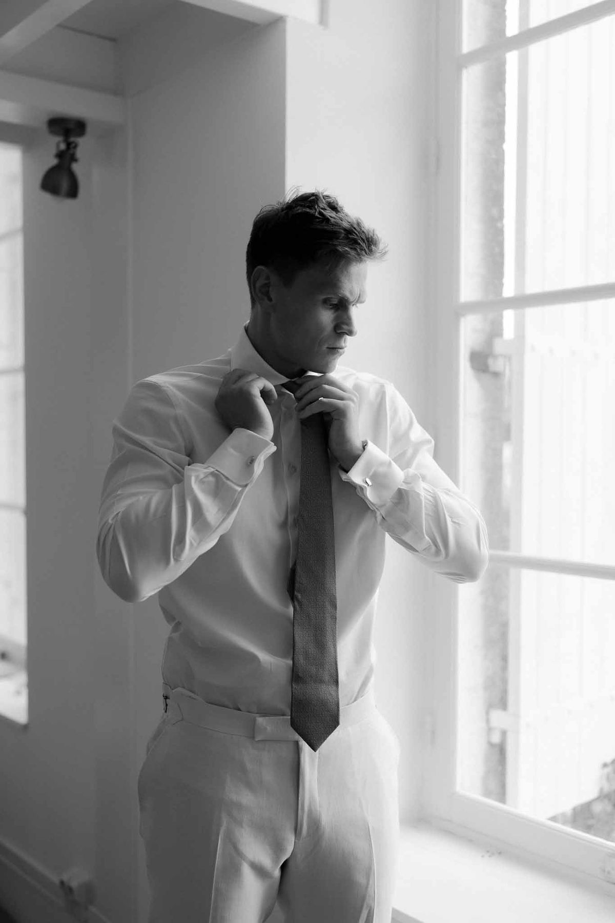 Black and white photo of groom adjusting tie by window during wedding morning preparations