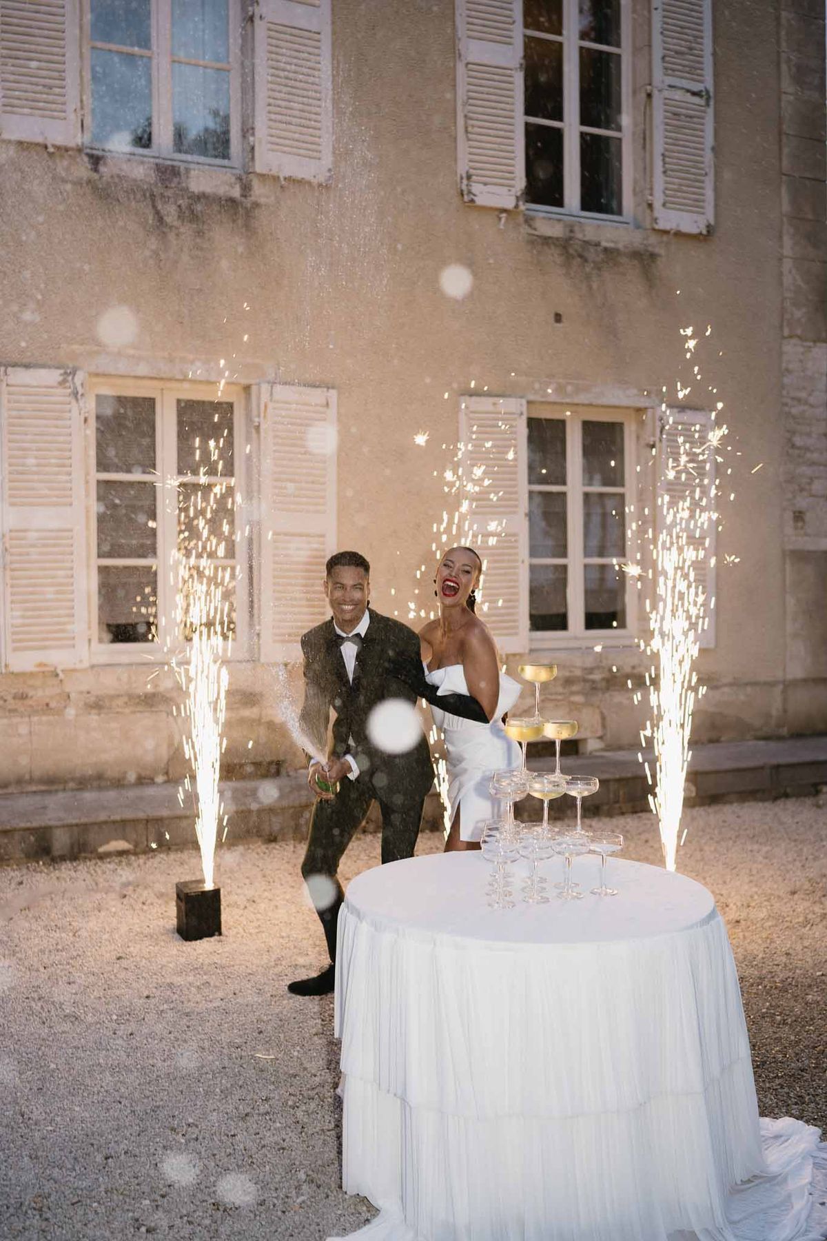 Couple celebrating with confetti during cocktail hour in historic stone courtyard with fairy lights and champagne tower