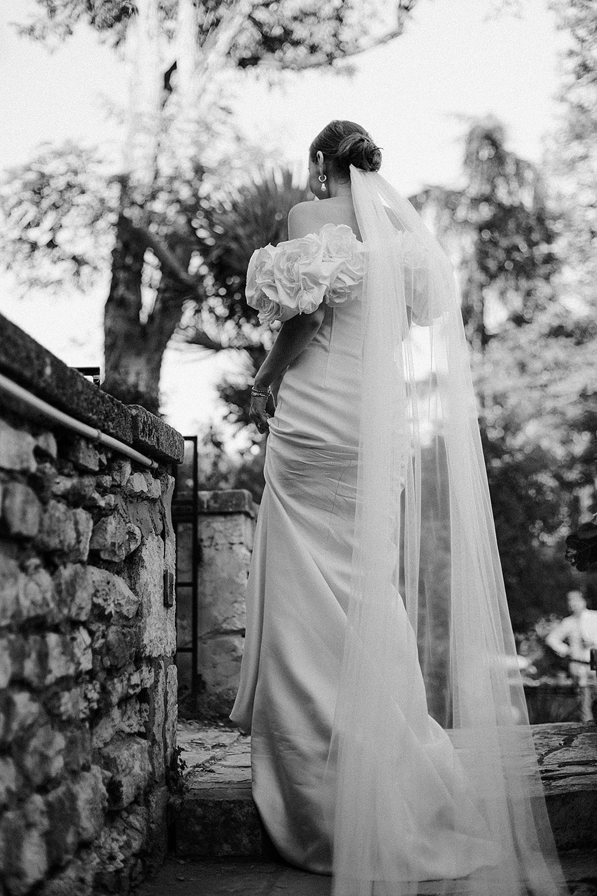 Bride in wedding gown with cathedral veil and bouquet in outdoor garden setting with stone wall