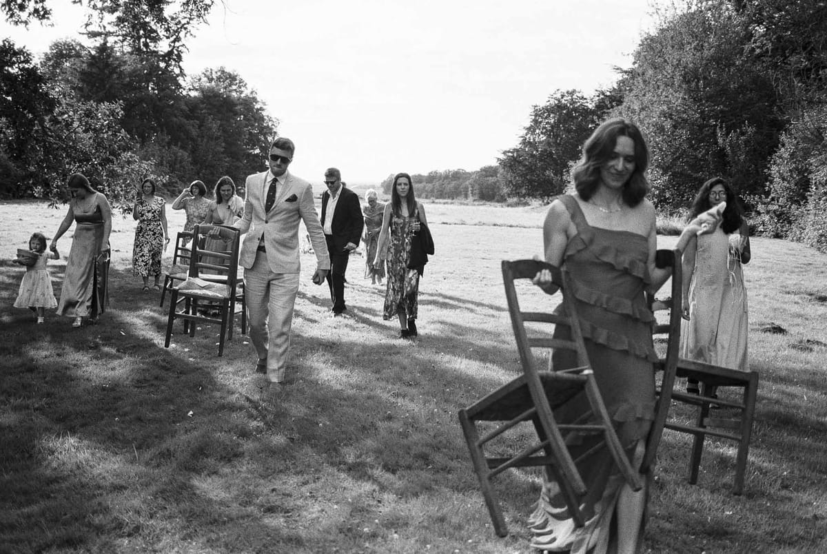 Black and white wide shot of outdoor wedding ceremony with guests scattered across riverside field