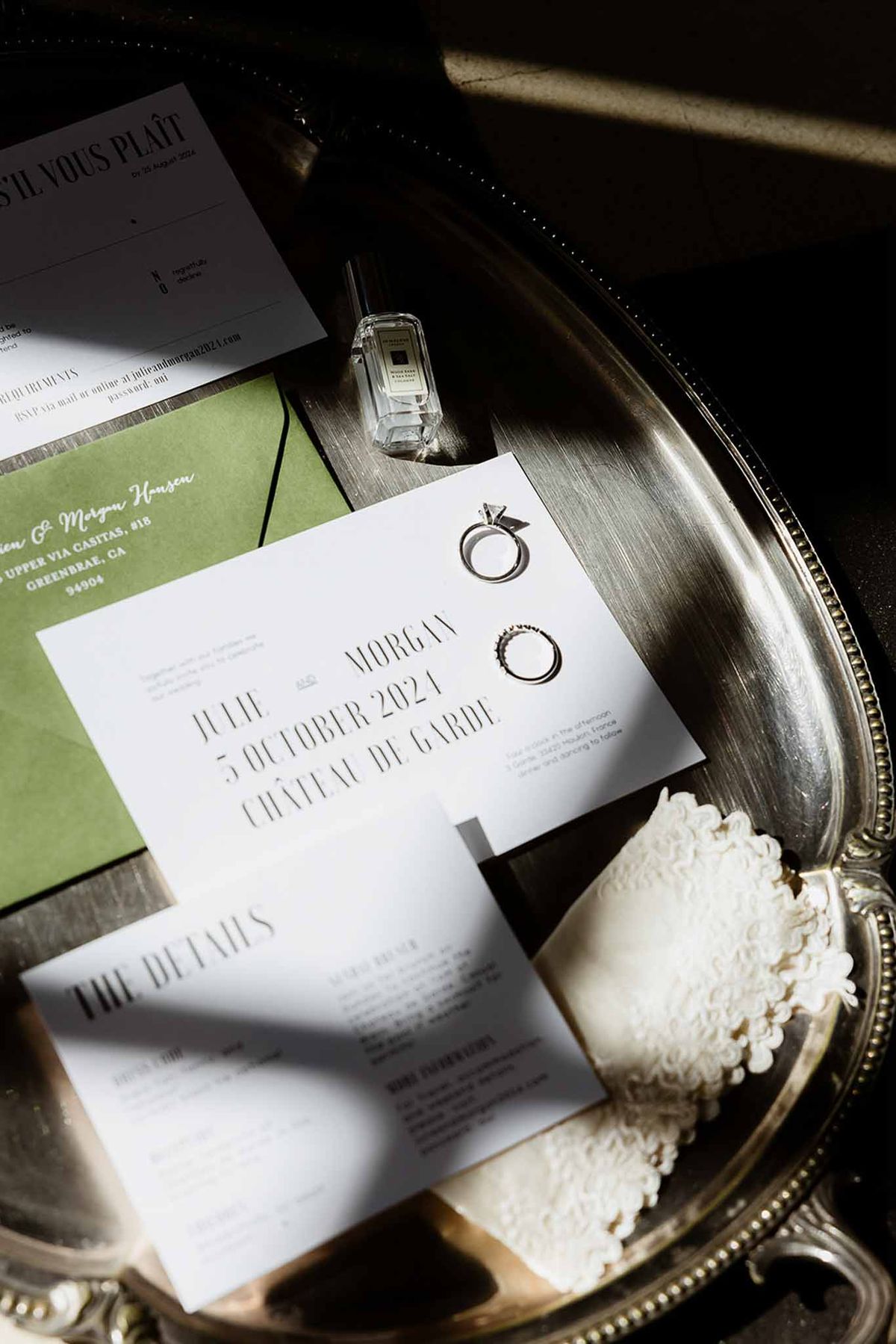 Wedding stationery flat lay with black serif typography, olive envelope, rings, and Jo Malone perfume