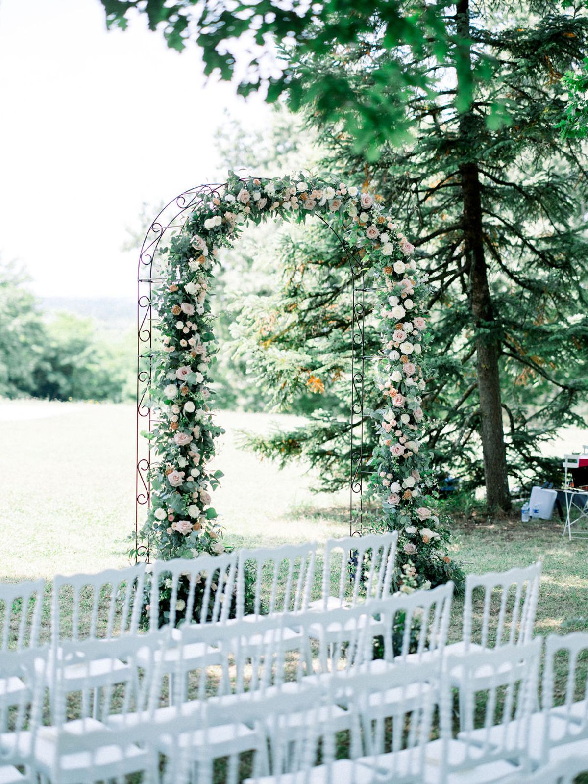 Wrought iron ceremony arch with blush roses and ivory blooms before semicircle of white Chiavari chairs