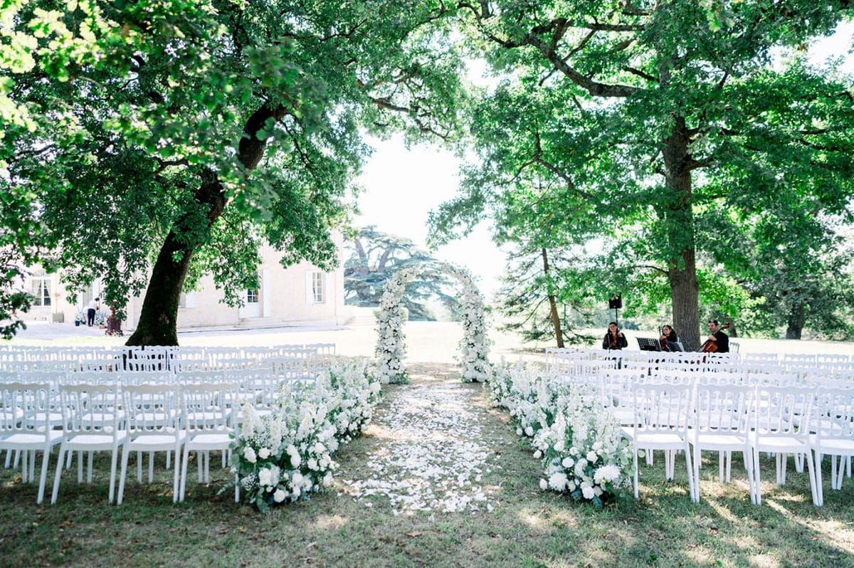 All-white ceremony setup with rose-covered arch, scattered petals, and string ensemble at chateau