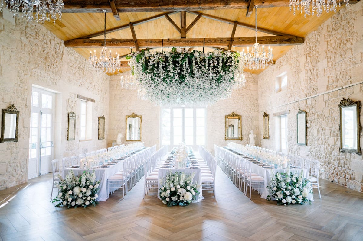 Three banquet tables with suspended white floral cloud and crystal chandeliers in vaulted stone mas with gold mirrors