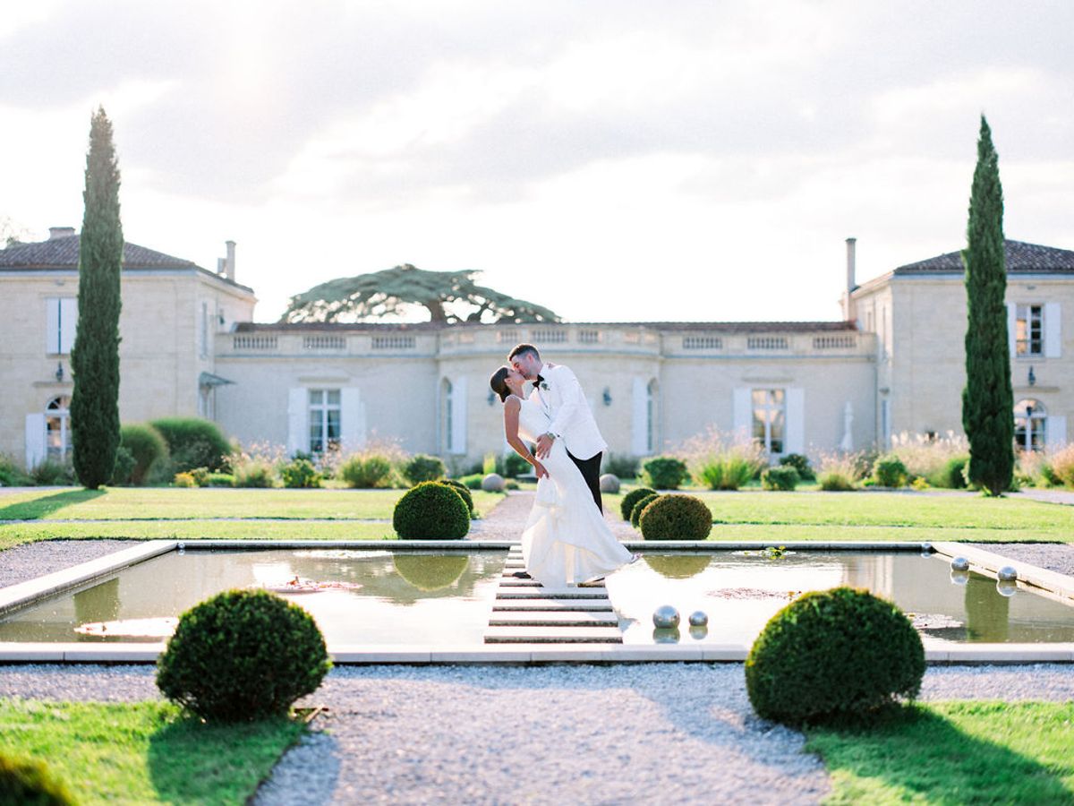 Bride and groom kiss on stone bridge over reflecting pool with chateau, cypress trees, and topiary beyond