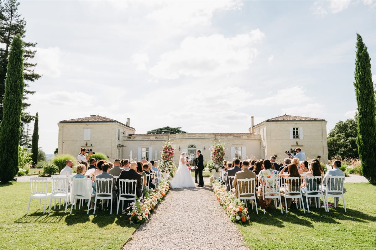 Outdoor chateau ceremony with couple at altar flanked by pink and coral floral installations and seated guests in white ch...